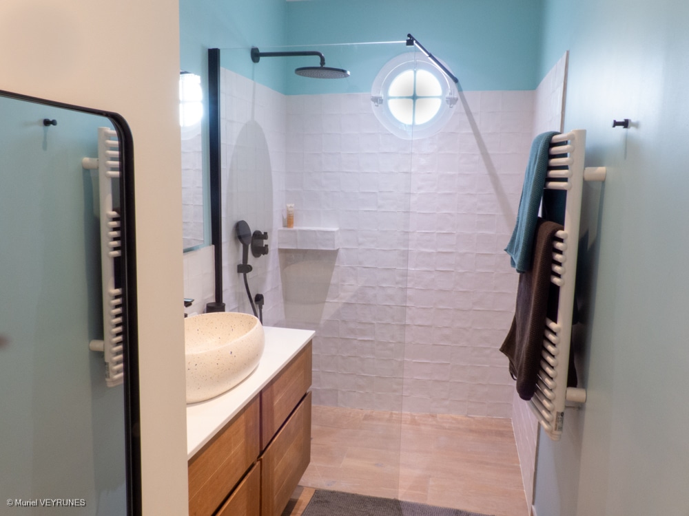 Salle de bain moderne et lumineuse : douche carrelage blanc, fenêtre ronde, vasque sur meuble bois, sèche-serviettes avec serviettes.