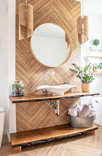 Salle de bain moderne avec mur bois style herringbone, miroir rond, vasque blanche, plan de travail et etagere en bois bord brut.