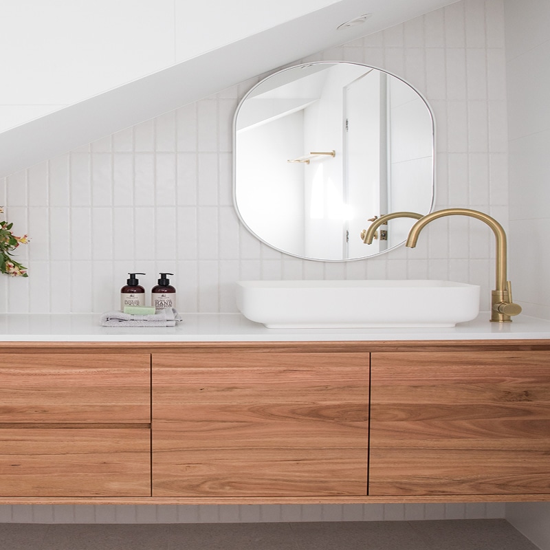 Salle de bain moderne avec meuble bois flottant, lavabo blanc, robinet doré, miroir ovale. Murs carrelés blanc. Bouteilles BODY WASH / HAND WASH.
