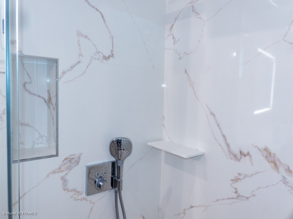 Douche moderne avec carrelage mural effet marbre blanc veiné de brun, robinetterie chromée, niche et étagère.