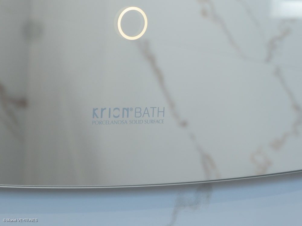Logo Krion® Bath Porcelanosa Solid Surface sur une surface blanche marbrée, avec un éclairage circulaire en haut.