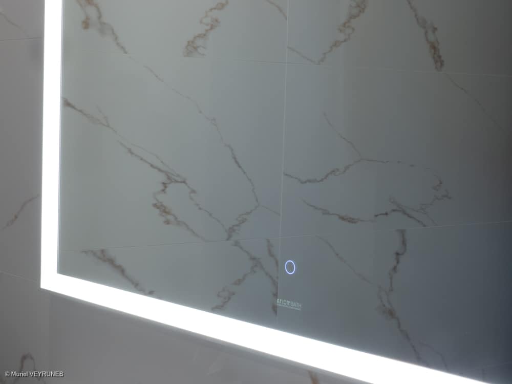 Miroir de salle de bain lumineux à LED intégré, tactile, reflétant un mur de carrelage blanc marbré avec des veines couleur rouille. Texte visible : KRYSTAL BATH.