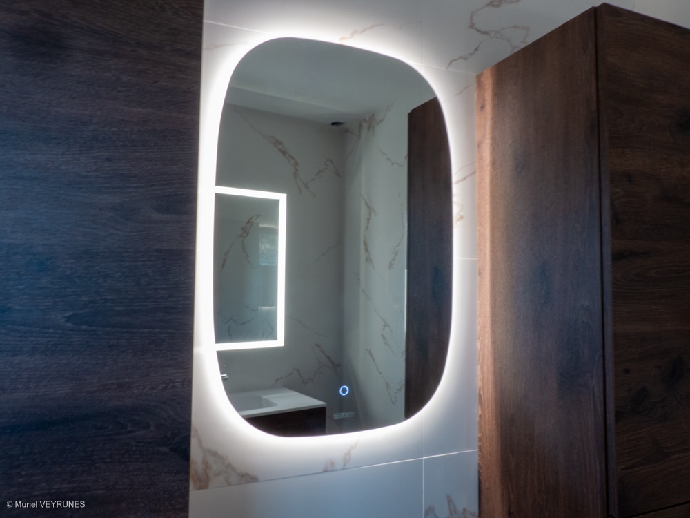 Miroir de salle de bain rétroéclairé à bords arrondis, reflétant un second miroir lumineux et un lavabo, encadré de bois sombre.