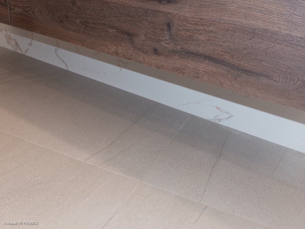 Plinthe effet marbre blanc veiné séparant un sol carrelé beige clair texturé d'un meuble bas imitation bois foncé.