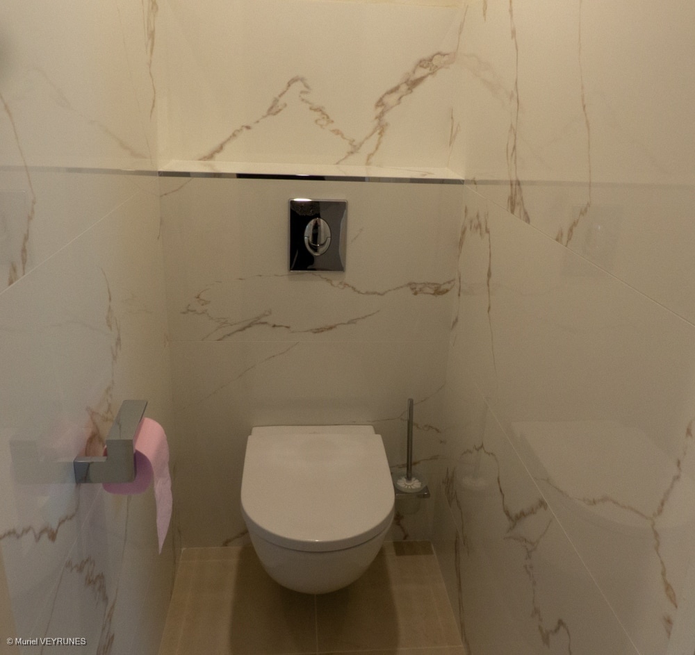 Toilettes suspendues modernes dans une salle d'eau carrelée blanc marbre, avec bouton poussoir chromé et porte-papier rose.